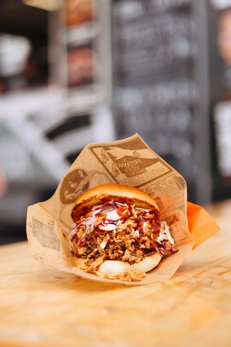 Un burger à la viande effilochée, chou rouge émincé, oignons frits et sauce barbecue, servi dans un papier kraft imprimé façon journal.